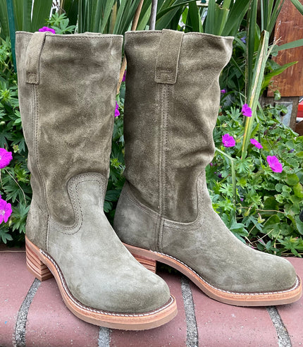 Frye green suede boots on a stone surface with plants and flowers in the background.