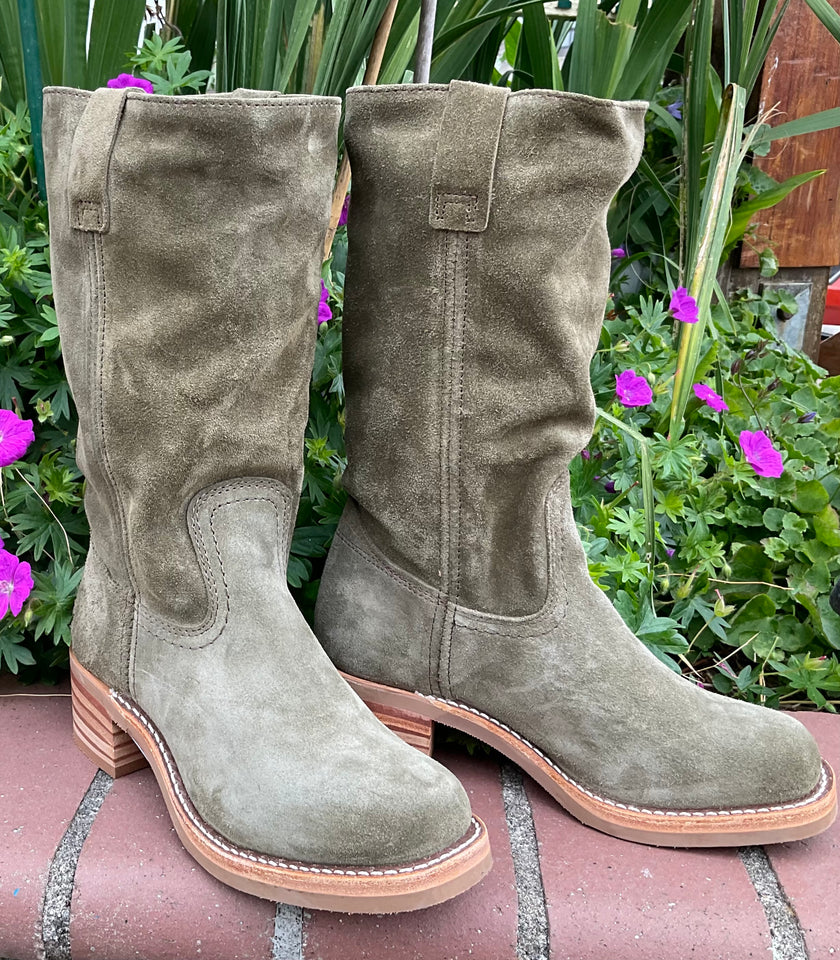 Frye green suede boots on a stone surface with plants and flowers in the background.