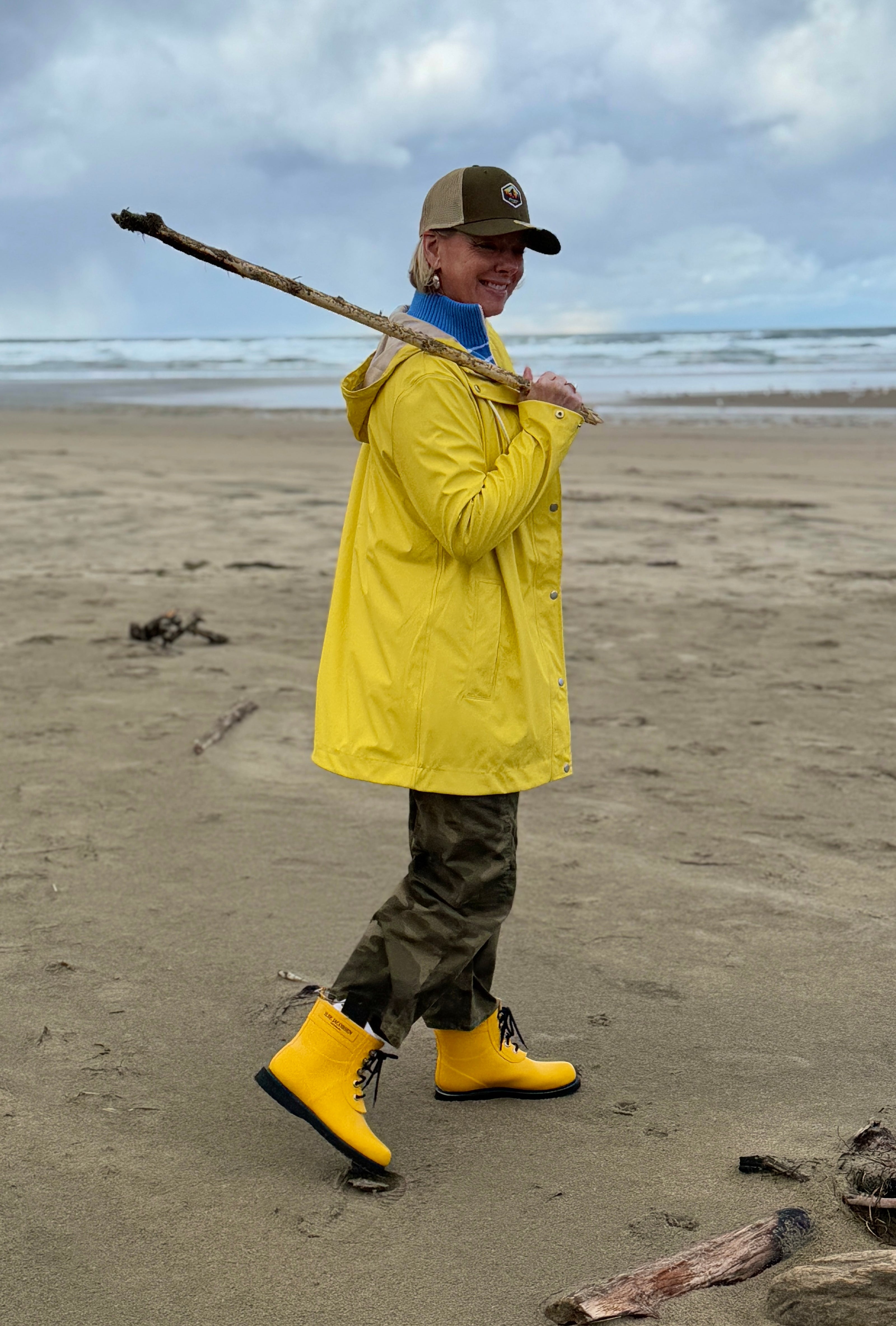 A person wearing a yellow rain jacket and boots, with a camouflage print on the lower half of the jacket, standing on a beach.
