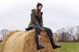 A person sitting on a hay bale wearing tall black rain boots with a decorative D-ring at the center back, paired with green pants.