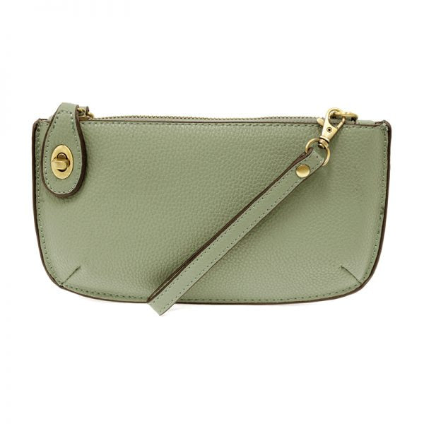 A sage-colored mini crossbody wristlet clutch with brass plated hardware and a printed lining, displayed with the wristlet strap visible