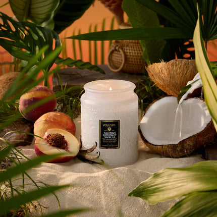 White candle with a label on a sandy surface surrounded by peaches and coconuts, with a blurred natural background.