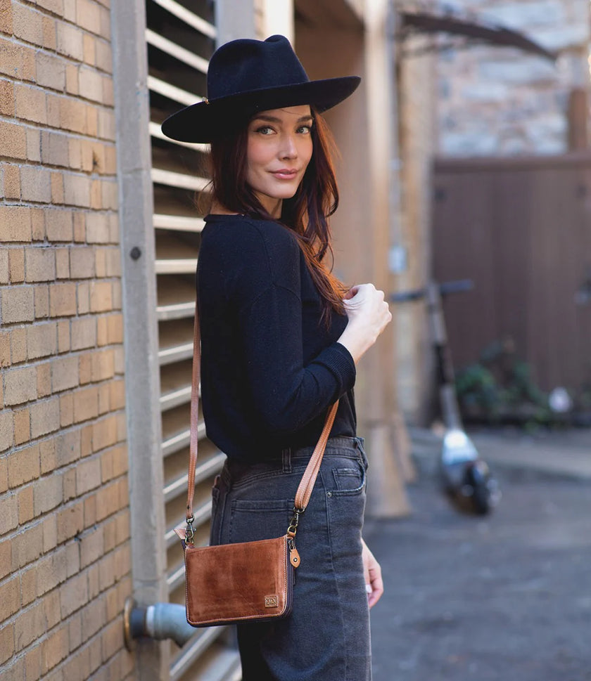 A woman with long brown hair in a black hat, black top, and gray jeans stands sideways in an alley, carrying the Bed|Stu Templeton ii Clutch Wallet crossbody. Brick walls and a blurred scooter are visible in the background.