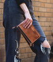 Wearing ripped black jeans and a dark sweater, a person holds the Bed|Stu Templeton ii Clutch Wallet—hand-painted leather with a zipper—while standing in front of a brick wall.
