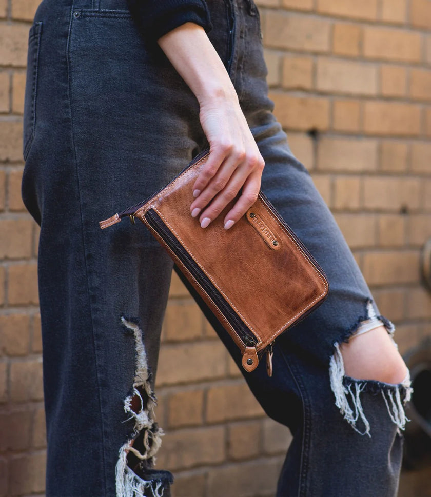 Wearing ripped black jeans and a dark sweater, a person holds the Bed|Stu Templeton ii Clutch Wallet—hand-painted leather with a zipper—while standing in front of a brick wall.