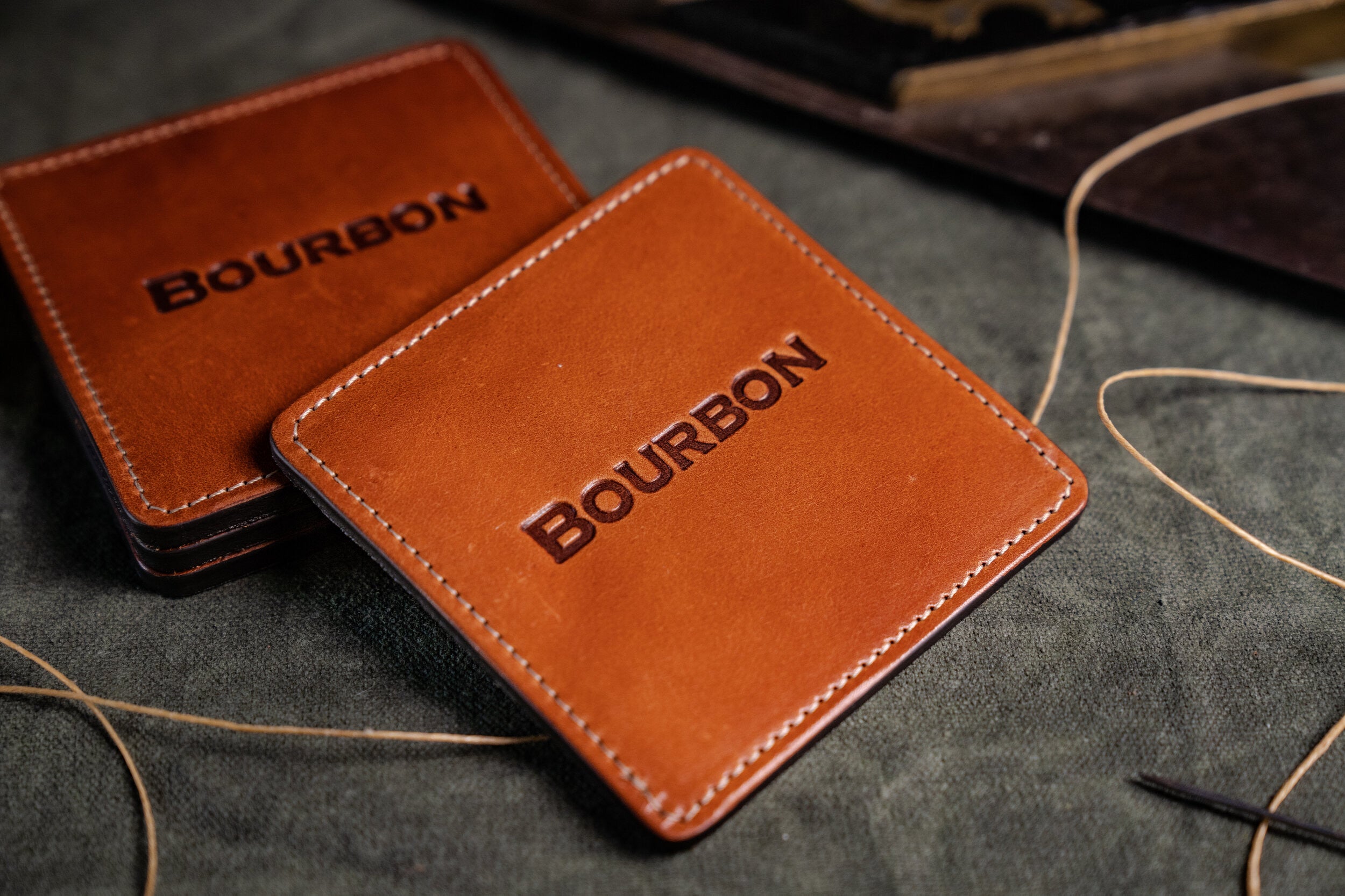 Two brown BourbonWare Classic Square Coasters, made from vegetable-tanned leather and embossed with BOURBON, rest on dark fabric beside stitching thread and a needle, reflecting their handcrafted quality.