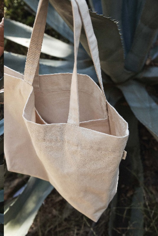 The Cheers Canvas Tote Bag - Natural, made of beige cotton canvas with two handles, hangs outdoors before large, pointed green leaves. Sunlight casts soft shadows on the bag and foliage, hinting at its spacious inside pocket.