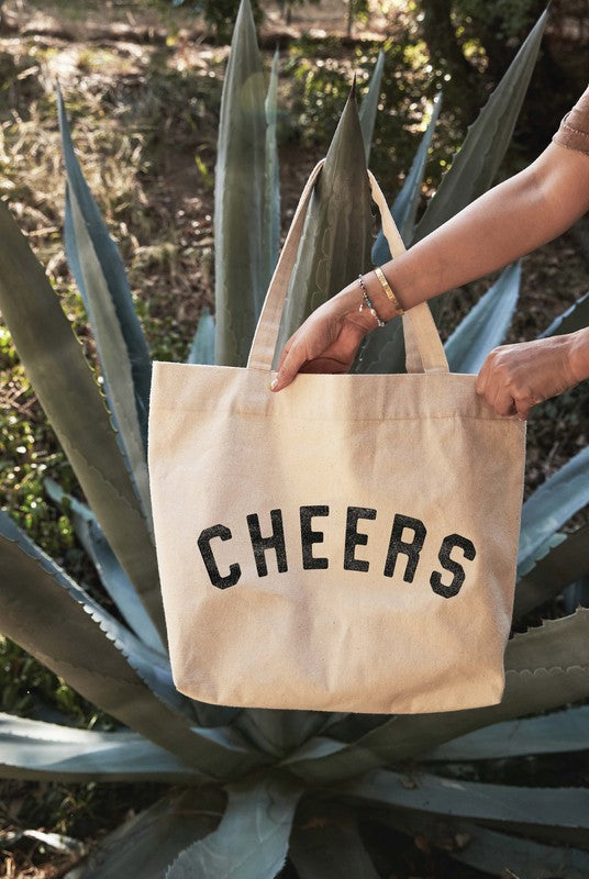 A person holds the Cheers Canvas Tote Bag - Natural, featuring bold black 